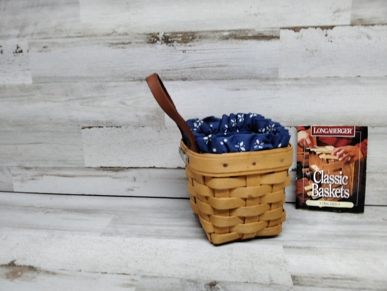 May include: A Longaberger Classic Baskets book with a woven basket with a blue and white fabric liner and a brown leather handle.