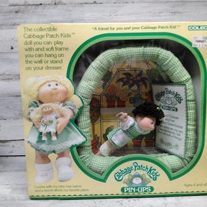 Puede incluir: Juego de muñecas coleccionables Cabbage Patch Kids Pin-Ups. El juego incluye dos muñecas, una que sostiene una muñeca más pequeña y una exhibición en forma de invernadero. El embalaje presenta texto y un patrón de cuadros verdes y blancos.