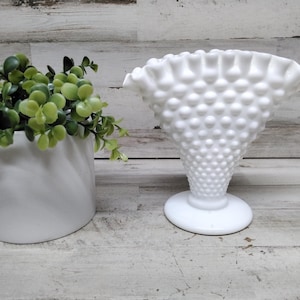 Puede incluir: Un jarrón blanco hobnail con borde con volantes y base redonda. El jarrón está junto a una maceta cilíndrica blanca con plantas verdes de hojas. Los objetos están sobre una superficie de madera clara.