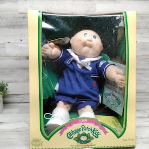 May include: A vintage Cabbage Patch Kids doll in its original packaging. The doll is wearing a blue sailor outfit with white accents and shoes. The packaging features the Cabbage Patch Kids logo and the year 1985.