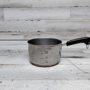 May include: A silver measuring cup with a black handle. The cup has markings for 1 cup, 3/4 cup, 1/2 cup, and 1/4 cup.