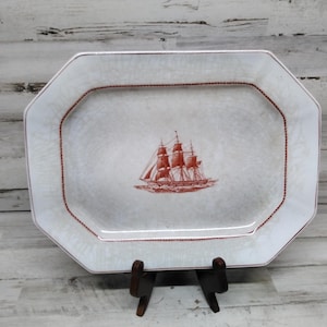 Puede incluir: Una fuente rectangular blanquecina con un borde rojo y una ilustración detallada de un velero. La fuente está sostenida por un soporte de madera, mostrando un tema náutico.