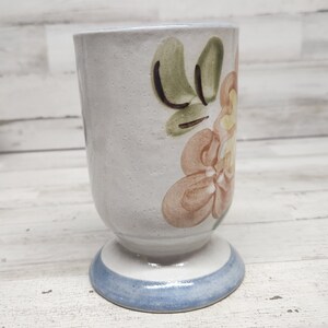 May include: A white ceramic mug with a floral design. The mug has a blue rim and a brown floral pattern with green leaves.