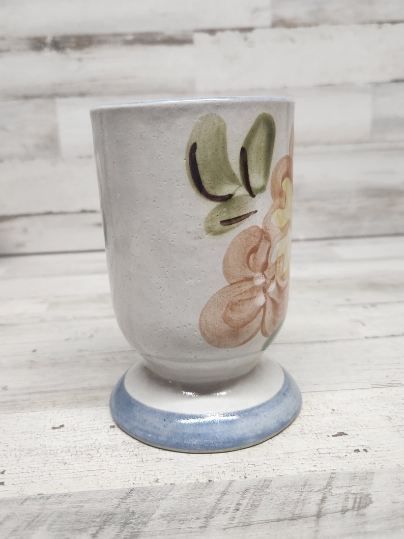 May include: A white ceramic mug with a floral design. The mug has a blue rim and a brown floral pattern with green leaves.