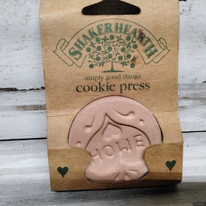 Puede incluir: Prensa para galletas rosa con forma de casa con la palabra «HOME» en relieve, en un embalaje de cartón marrón. El embalaje lleva las palabras «SHAKER HEARTH» y «cookie press».
