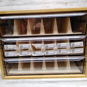 May include: A wooden storage unit with three clear plastic drawers. Each drawer contains multiple items, some of which are labeled with the number "29" and the word "FLOSS". The unit is made of light-colored wood.