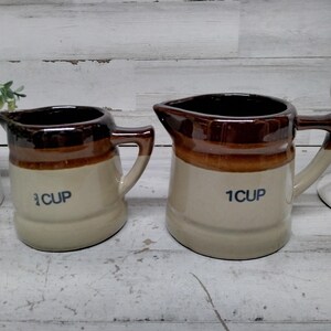 Op de afbeelding: Twee keramische maatbekers. Eén is gemarkeerd met "1/3 CUP" en de andere met "1 CUP". Beide hebben een bruine rand en handvat, met een beige body. De bekers zijn ontworpen met een schenktuit.