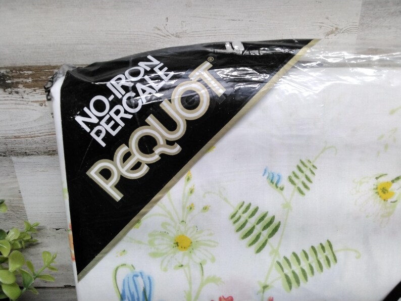 K&ouml;nnte beinhalten: Eine Packung wei&szlig;en Perkalstoffs mit floralem Muster. Die Packung hat ein schwarzes Etikett mit den wei&szlig;en Worten "NO-IRON PERCALE PEQUOT". Das florale Muster umfasst G&auml;nsebl&uuml;mchen, Glockenblumen und gr&uuml;ne Bl&auml;tter.