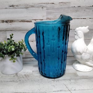 May include: A teal glass jug with a textured design and a handle. A white ceramic cockerel ornament and a small potted plant are also present. The jug is a decorative item, suitable for serving drinks.