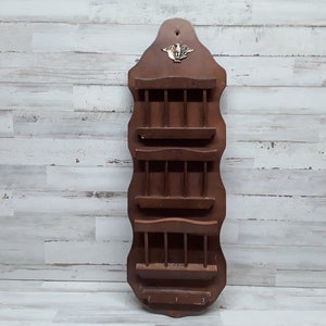 May include: A vintage wooden wall-mounted organizer with multiple shelves and a key rack. The dark brown organizer features a decorative brass emblem at the top and is designed for mail or small items. The piece is against a white and gray wood-paneled wall.