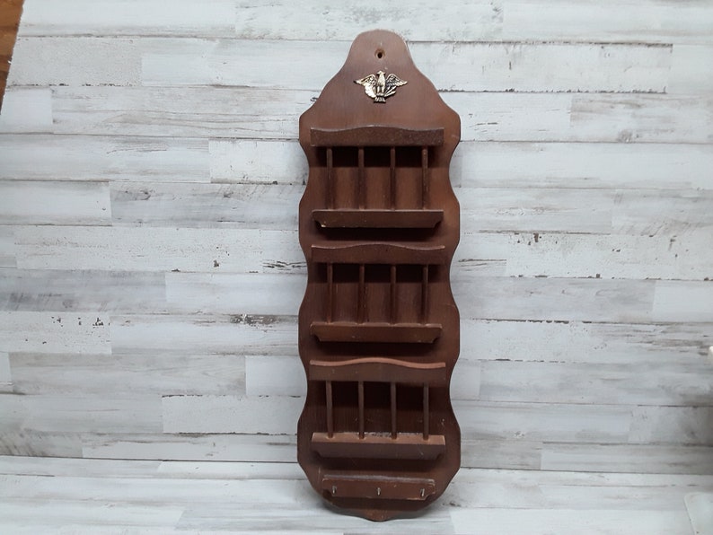 May include: A vintage wooden wall-mounted organizer with multiple shelves and a key rack. The dark brown organizer features a decorative brass emblem at the top and is designed for mail or small items. The piece is against a white and gray wood-paneled wall.