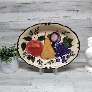 以下が含まれることがあります： 絵付けされたフルーツのデザインが施された楕円形のセラミック製サービングプラッター。赤いリンゴ、黄色の梨、紫色のプラム、ブドウの房が特徴です。縁はスカラップで、濃い緑色の縁取りが施されています。白いセラミック製の雄鶏が背景にあります。