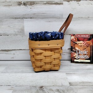 May include: A small, brown wicker basket with a blue and white fabric liner and a brown leather handle. The basket is sitting on a white wood surface. A small book titled "Longaberger Classic Baskets" is sitting next to the basket.