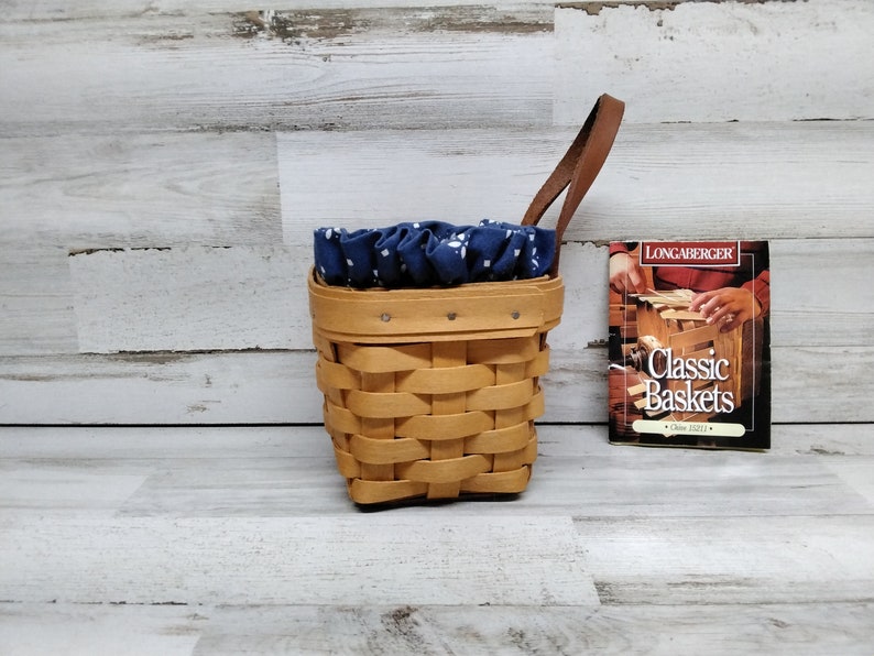 May include: A small, brown wicker basket with a blue and white fabric liner and a brown leather handle. The basket is sitting on a white wood surface. A small book titled "Longaberger Classic Baskets" is sitting next to the basket.
