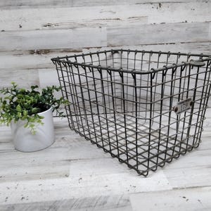 Könnte beinhalten: Ein rechteckiger, dunkelgrauer Drahtkorb mit einem kleinen Metallschild. Der Korb steht neben einem weißen Keramiktopf mit grünen Pflanzen. Der Hintergrund ist eine weiß-graue Holzpaneelwand.