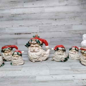 Könnte beinhalten: Eine Sammlung von Keramik-Weihnachtsmannfiguren und eine weiße Hahnfigur. Die Weihnachtsmannfiguren haben rote Mützen und weiße Bärte. Die Weihnachtsmannfiguren variieren in Größe und Form, ideal für die Weihnachtsdekoration.