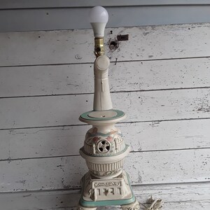 May include: A white ceramic lamp shaped like a vintage wood-burning stove with a green base and floral accents. The lamp has a light bulb and a cord.