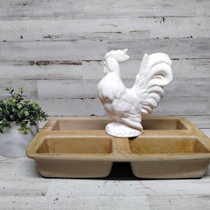 Puede incluir: Una figura de gallo de cerámica blanca se encuentra sobre una fuente rectangular beige dividida en cuatro compartimentos. A la izquierda de la fuente hay una pequeña planta en maceta con hojas verdes.