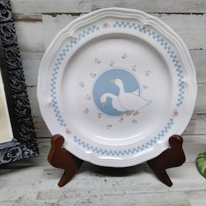 May include: White ceramic plate with a blue and white checkered border and a central design featuring two white geese in a blue circle. The plate is displayed on a brown wooden stand, with a decorative frame and a goose figurine in the background.