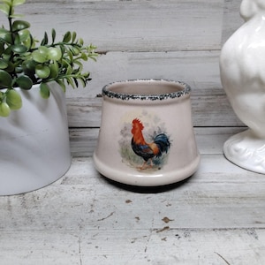 Puede incluir: Un portautensilios de cerámica con una ilustración de gallo. El portautensilios es de color blanquecino con un borde verde oscuro y azul. El gallo está representado con plumas rojas, azules y marrones. También se ve una figura de gallo de cerámica blanca.