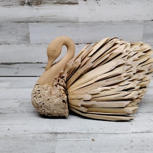May include: A wooden swan figurine with a brown, straw-like tail. The swan's head is turned to the side, and its beak is closed. The figurine is made of natural materials and has a rustic look.
