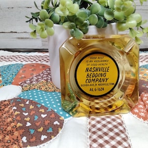 May include: A vintage amber-colored glass ashtray with the text "NASHVILLE BEDDING COMPANY" in a yellow circle. The ashtray sits on a colorful patchwork quilt, with a white vase and green plant in the background.