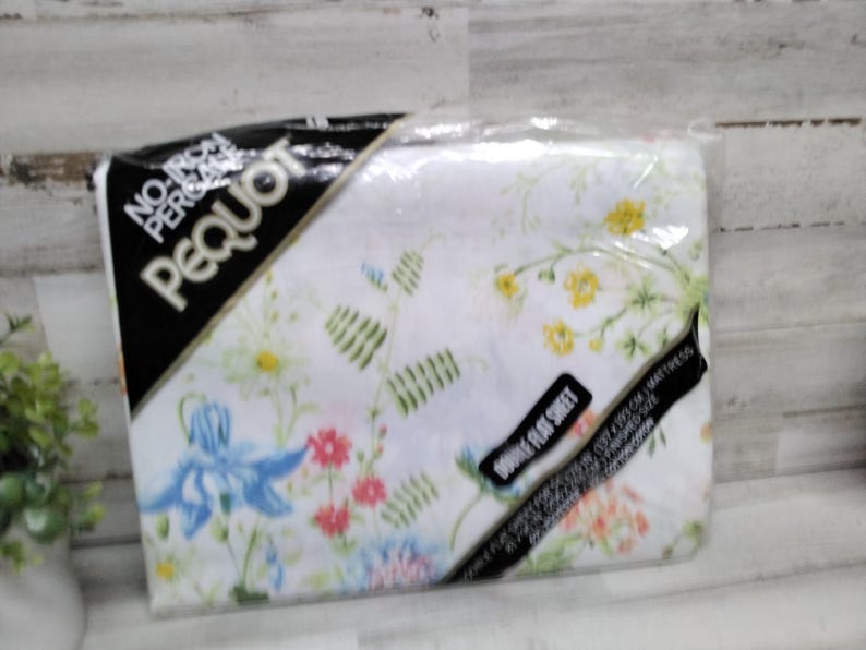 K&ouml;nnte beinhalten: Ein verpacktes Doppel-Flachbettlaken mit floralem Muster. Das Laken ist wei&szlig; mit blauen, rosa und gelben Blumen und gr&uuml;nen Bl&auml;ttern. Die Verpackung tr&auml;gt den Markennamen "Pequot" und die Worte "No Iron, Perma-Press".