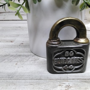 May include: A small, antique-style padlock with a dark grey body and a gold-coloured handle. The text "BG BLUEGRASS 44" is embossed on the front. The padlock is likely a decorative item or collectible.