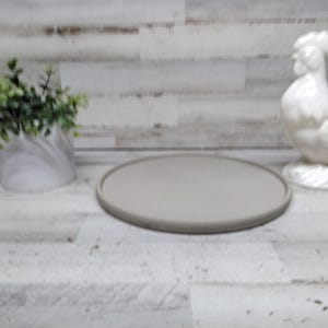 May include: A round, gray serving tray with a raised edge sits on a light-colored wooden surface. A white ceramic rooster figurine and a small potted plant with green foliage are also present.