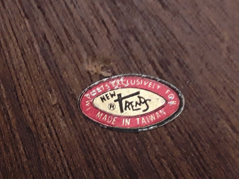 May include: A close-up of a label with the text "NEW TRENDS" in a stylized font, surrounded by a red oval border. The label also includes the text "MADE IN TAIWAN". The background is a dark wood grain.