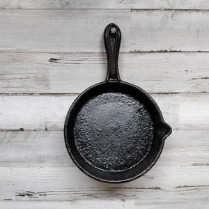 May include: A black cast iron skillet with a long handle sits on a white wooden surface.