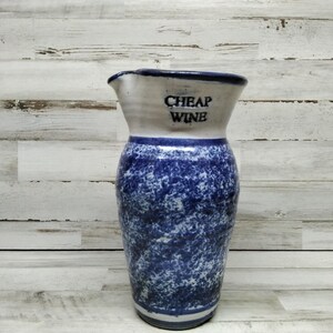 May include: A ceramic pitcher with a white rim and a blue speckled body. The words "CHEAP WINE" are printed in black on the white section. The pitcher has a spout for pouring.