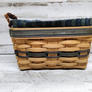 May include: Woven rectangular basket in light brown and dark green. The basket features a woven design with a dark green band and trim. The top edge is lined with dark green and blue fabric. The handles are made of twisted brown material.