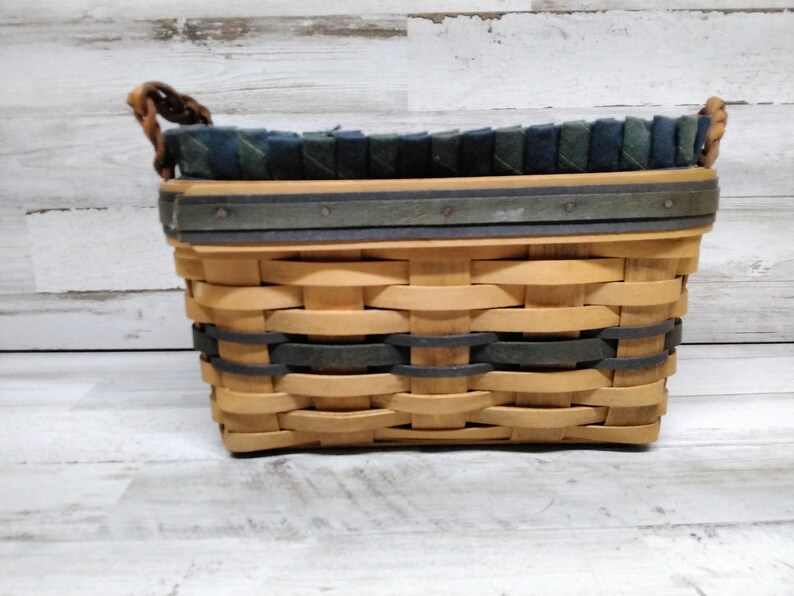May include: Woven rectangular basket in light brown and dark green. The basket features a woven design with a dark green band and trim. The top edge is lined with dark green and blue fabric. The handles are made of twisted brown material.