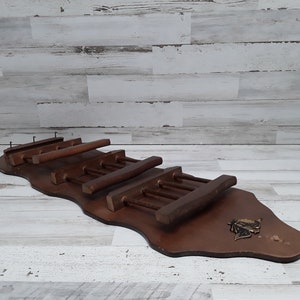 May include: A dark brown wooden wall-mounted wine rack with a decorative metal emblem. The rack features multiple horizontal slots for holding wine bottles and glasses. The rack is designed to hold wine bottles and glasses.