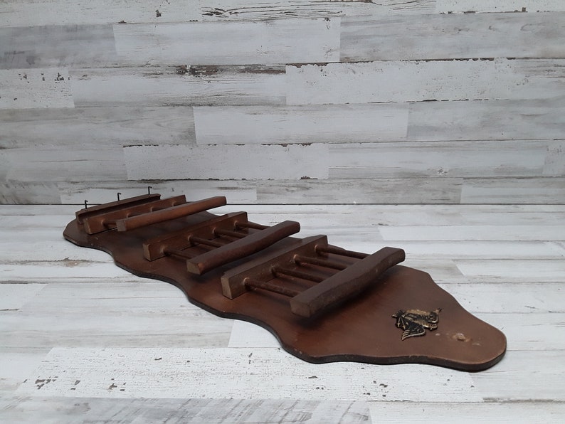 May include: A dark brown wooden wall-mounted wine rack with a decorative metal emblem. The rack features multiple horizontal slots for holding wine bottles and glasses. The rack is designed to hold wine bottles and glasses.