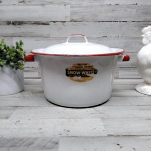 May include: White enamel pot with a red rim and handles, and a matching lid. The pot has a label that reads "Snow White". A white ceramic rooster and a small white pot with greenery are also in the image.