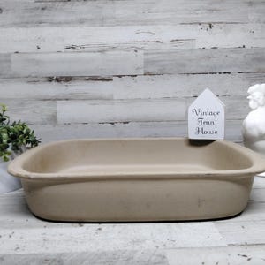 May include: A rectangular, light beige stoneware baking dish with rounded corners and a smooth, matte finish. The dish features a wide, flat rim and deep sides, presenting a neutral, unglazed appearance suitable for oven-to-table food preparation.