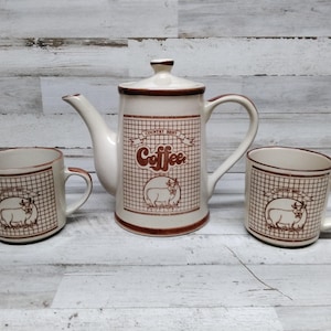 May include: A white ceramic coffee pot with a brown trim and a cow design. The pot says "Country Roast Coffee" on the front. Two matching mugs with the same design are on either side of the pot.