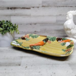 Puede incluir: Una fuente rectangular de cerámica amarilla con detalles de zanahorias naranjas y hojas verdes. Un gallo de cerámica blanca y una pequeña maceta blanca con follaje verde están en el fondo.