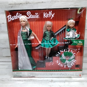 Op de afbeelding: Een Barbie, Stacie en Kelly poppenset in een doos. De poppen dragen groene en zilveren feestjurken. De doos heeft de tekst "Singing Holiday Sisters" en "Try Me".