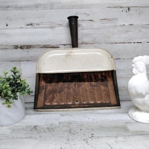 Puede incluir: Un recogedor de metal vintage con una sección superior de color crema y una base interior de color marrón oscuro oxidado con un mango vertical. El recogedor se encuentra entre una pequeña maceta blanca con follaje verde y una figura de gallo.