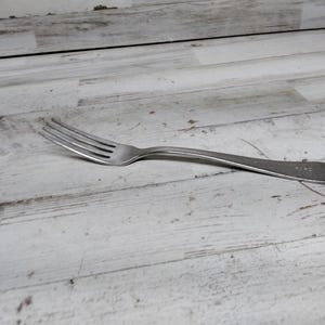 May include: A silver fork with a simple design. The fork is lying on a white wooden surface.