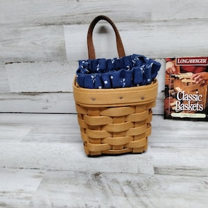 May include: A small, brown wicker basket with a blue and white fabric liner. The basket has a leather handle and is hanging on a wall. A small book titled "Classic Baskets" is visible in the background.