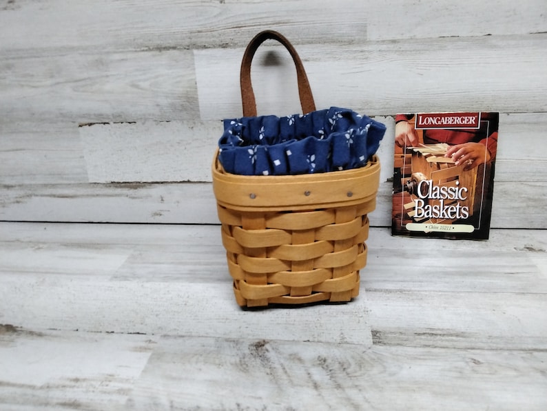 May include: A small, brown wicker basket with a blue and white fabric liner. The basket has a leather handle and is hanging on a wall. A small book titled "Classic Baskets" is visible in the background.