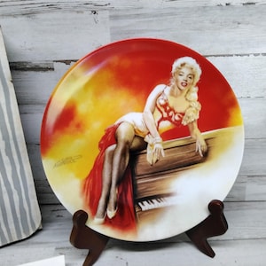 Puede incluir: Un plato de cerámica blanco con un diseño rojo, amarillo y blanco que presenta a una mujer con un vestido rojo sentada en un piano. El plato está en un soporte de madera.