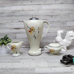 Puede incluir: Un juego vintage de cafetera eléctrica, crema y azucarero de color crema con detalles florales. También hay una figura de gallo de cerámica blanca y una pequeña planta en maceta. Un cable eléctrico negro está en primer plano.