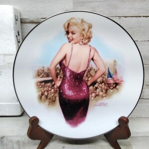 Puede incluir: Un plato de cerámica blanco con un borde negro presenta una ilustración colorida de una mujer con un vestido rojo de lentejuelas. La mujer está de pie frente a una multitud de personas y una bandera estadounidense.