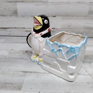 Puede incluir: Un macetero de cerámica con forma de pingüino con cuerpo blanco, alas negras y pico amarillo. El pingüino lleva una bufanda roja y blanca. El macetero está sobre una superficie de madera blanca. El pingüino sostiene un macetero de cerámica blanco con forma de cubo de hielo con detalles azules.