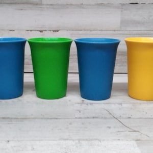 Puede incluir: Cuatro vasos de plástico de diferentes colores: azul, verde, azul y amarillo. Los vasos están alineados en una fila sobre una superficie de madera blanca.